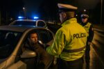 Dvaja slovenskí policajti v reflexných vestách s nápisom "POLÍCIA" zastavili osobné auto na nočnej ceste. Jeden z policajtov dáva vodičovi fúkať do alkohol testera, zatiaľ čo druhý policajt stojí opodiaľ. V pozadí je vidieť ďalšie policajné vozidlo s rozsvietenými majákmi.