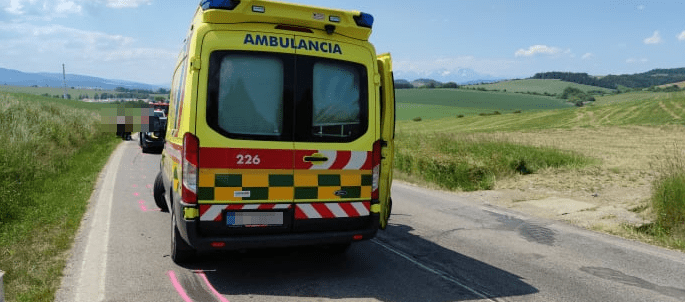 Žltá sanitka s evidenčným číslom 226 stojí na ceste II/533 medzi Levočou a Spišskou Novou Vesou. Záchranárske vozidlo je zaparkované na okraji vozovky s otvorenými zadnými dverami. V pozadí vidno poľnohospodársku krajinu, trávnaté porasty a vzdialené pohorie Vysokých Tatier pod modrou oblohou. Na asfalte sú viditeľné ružové značky vyznačujúce miesto dopravnej nehody. (Obrázok ilustruje zásah záchranárov pri udalosti na Spiši).