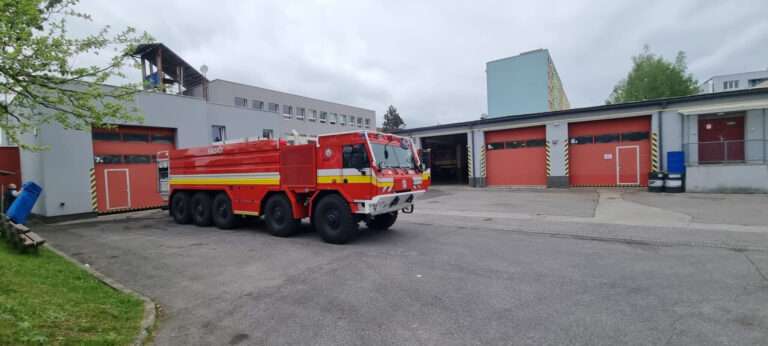 Pohľad na výkonné červené hasičské cisternové vozidlo typu 8x8 s nápisom „HASIČI“ a reflexnými pásmi, zaparkované na asfaltovom nádvorí hasičskej stanice s viacerými garážovými bránami, pod oblačnou oblohou.