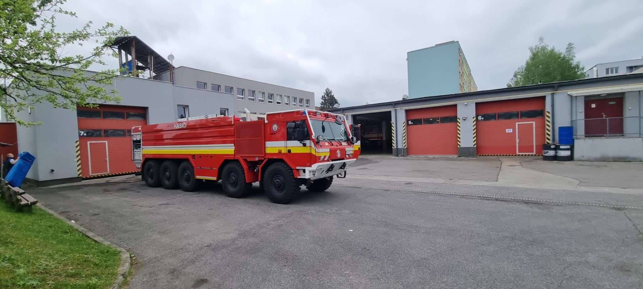 Pohľad na výkonné červené hasičské cisternové vozidlo typu 8x8 s nápisom „HASIČI“ a reflexnými pásmi, zaparkované na asfaltovom nádvorí hasičskej stanice s viacerými garážovými bránami, pod oblačnou oblohou.