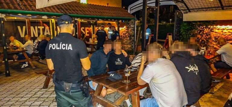 Nočná kontrola polície na letnej terase reštaurácie. Policajt s nápisom POLÍCIA na chrbte stojí pri drevenom stole, kde sedí skupina mladých ľudí s pohármi piva. Tváre civilistov sú rozmazané.