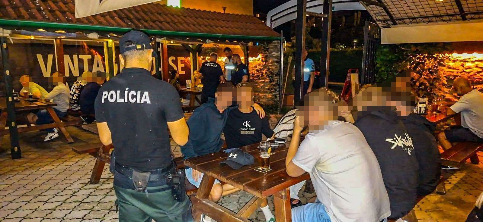 Nočná kontrola polície na letnej terase reštaurácie. Policajt s nápisom POLÍCIA na chrbte stojí pri drevenom stole, kde sedí skupina mladých ľudí s pohármi piva. Tváre civilistov sú rozmazané.