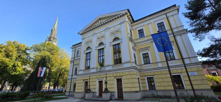 Historická budova radnice v centre Spišskej Novej Vsi počas slnečného dňa s kostolnou vežou v pozadí. Sídlo mestského úradu a mestskej volebnej komisie pre komunálne voľby 2026.