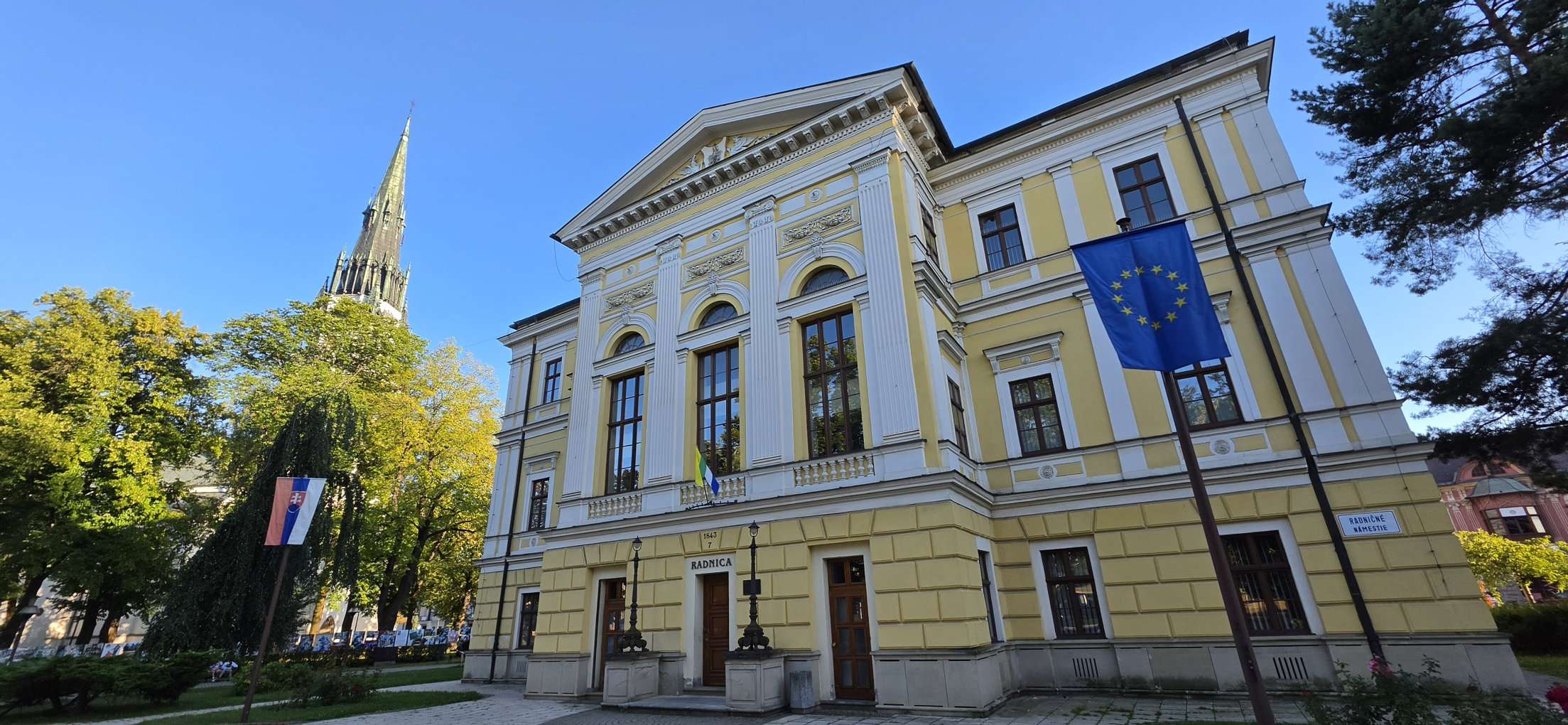 Historická budova radnice v centre Spišskej Novej Vsi počas slnečného dňa s kostolnou vežou v pozadí. Sídlo mestského úradu a mestskej volebnej komisie pre komunálne voľby 2026.