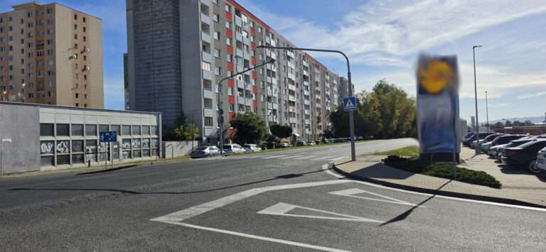 Pohľad z úrovne ulice na širokú mestskú cestu na sídlisku v slnečný deň. V popredí sú na asfalte výrazné biele vodorovné dopravné značenie v tvare šípok smerujúcich vpred. Vľavo stojí nízkopodlažná komerčná budova s oknami a modrým nápisom, vedľa nej sa tiahne dlhý panelový bytový dom so sivou a červenou fasádou. Napravo je na stĺpe umiestnený rozmazaný (cenzurovaný) komerčný nápis (pravdepodobne logo obchodu) vedľa veľkého parkoviska plného áut pri križovatke. Za cestou sa dvíha ďalší veľmi vysoký panelový obytný dom. Scénu lemujú ďalšie panelové bytové domy a roztrúsená zeleň stromov pod jasnou modrou oblohou s rozptýlenými oblakmi. Pozdĺž trasy sú viditeľné pouličné osvetlenie a dopravné značky. V diaľke sa na horizonte črtá mestská zástavba. Perspektíva je z pohľadu vodiča alebo chodca z boku cesty