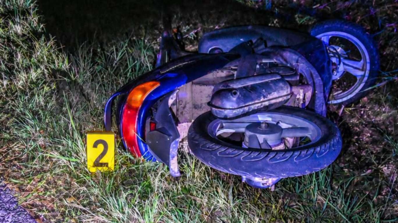Nočná fotografia prevráteného tmavomodrého skútra ležiaceho v tráve v priekope. Pri kapotáži je položený žltý policajný evidenčný terčík s číslom 2.