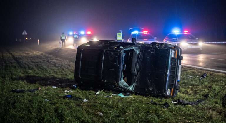 Nočný dokumentárny záber zblízka na silne poškodený, starší model tmavého Jeepu Cherokee, ktorý leží prevrátený na boku v trávnatom svahu vedľa cesty. Scéna je v hustom nočnom opare, osvetlená reflektormi záchranárov. V rozostrenom pozadí je vidieť blikajúce modré policajné majáky z viacerých policajných vozidiel a policajnú hliadku v reflexnej veste.