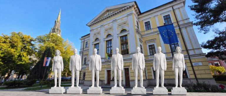 7 bielych sôch symbolizujúcich kandidátov na primátora pred budovou radnice. Ilustračná snímka pre komunálne voľby Spišská Nová Ves 2026