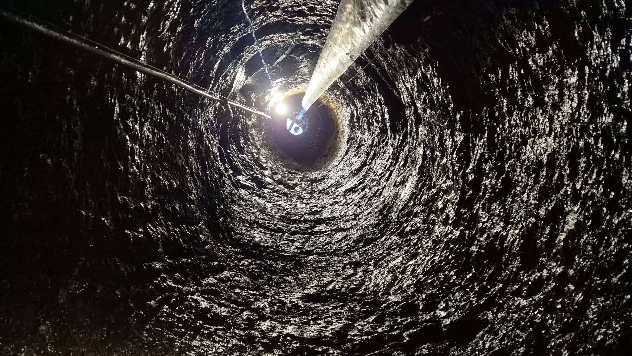 Dramatický pohľad zvnútra hlbokej, kruhovej skalnej šachty s drsnými, vlhkými stenami. Svetlo prichádza zhora z ústia a silné reflektory nižšie vytvárajú oslnenie a perspektívu hĺbky. Strieborná rúra a káble prechádzajú stredom.