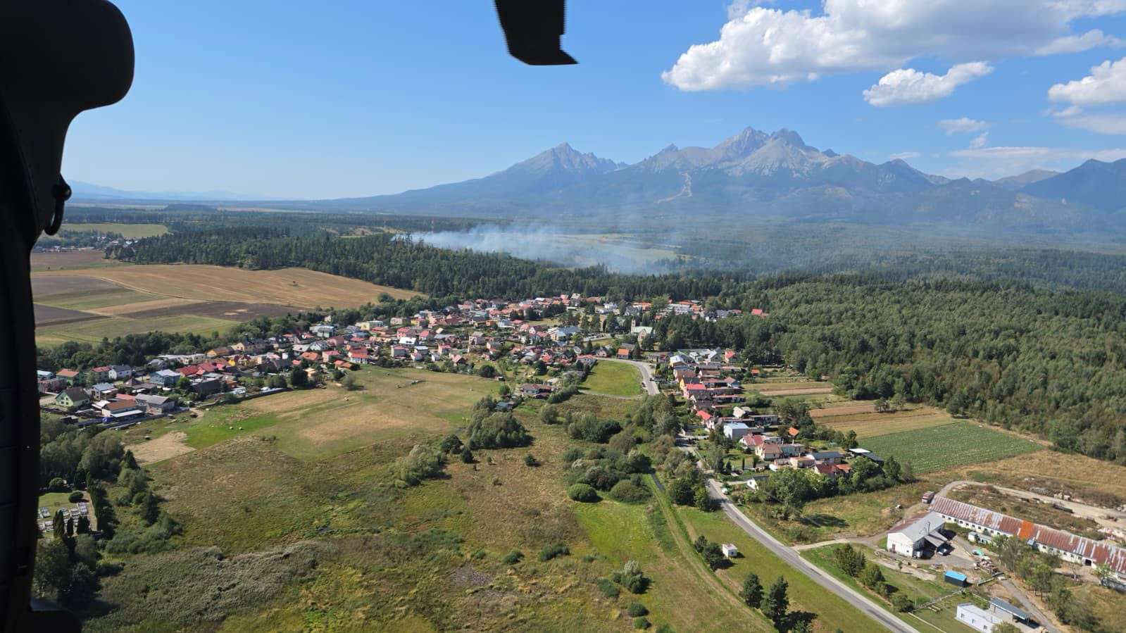 Letecký záber na podtatranskú obec z vrtuľníka počas zásahu pri požiari lesného porastu pri Stráňach pod Tatrami. V pozadí panoráma Vysokých Tatier s dymom nad lesom pod modrou oblohou.