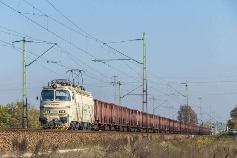 Nákladný vlak ťahaný svetlým elektrickým rušňom s dlhým radom hnedých vozňov prechádza po železničnej trati počas slnečného dňa.