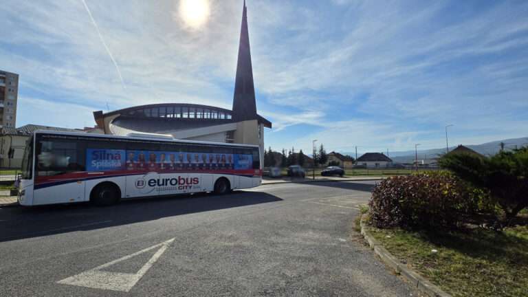 Fotografia bieleho autobusu MHD spoločnosti eurobus s celoplošným modrým reklamným polepom kandidátov klubu Silná Spišská na bočnej strane. Autobus stojí na ceste pred moderným kostolom počas slnečného dňa.