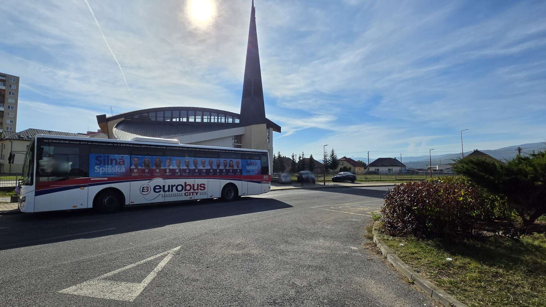 Fotografia bieleho autobusu MHD spoločnosti eurobus s celoplošným modrým reklamným polepom kandidátov klubu Silná Spišská na bočnej strane. Autobus stojí na ceste pred moderným kostolom počas slnečného dňa.