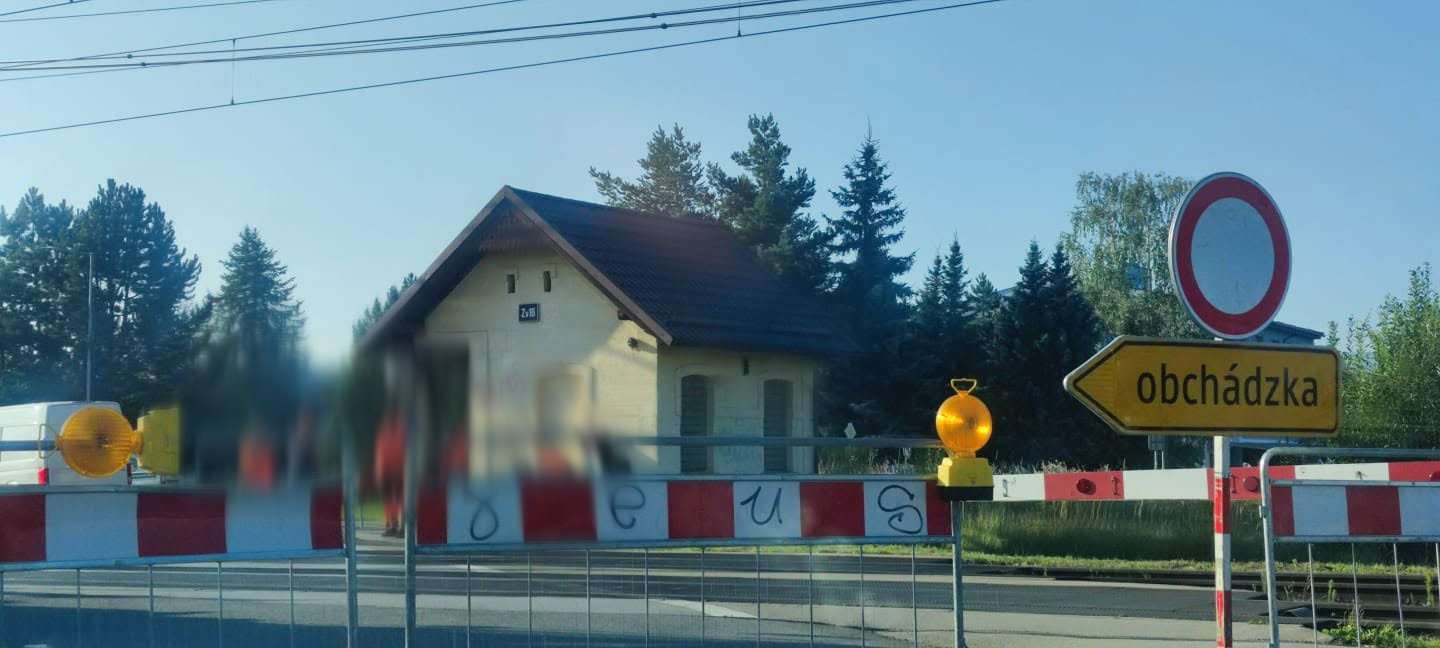 Dopravné značenie zákazu vjazdu a smerovej šípky s nápisom 'obchádzka' pred železničným priecestím. V pozadí je malá železničná budova a stromy.