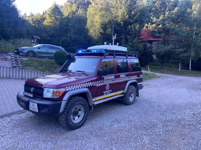 Bordové terénne vozidlo Toyota Land Cruiser Horskej záchrannej služby (HZS) s evidenčným číslom SN-839AY, so zapnutými modrými majákmi na streche, stojí na štrkovej prístupovej ceste. Na dverách vozidla je viditeľný nápis „ZÁCHRANNÉ VOZIDLO“ a logo HZS „SLOVENSKÝ RAJ“. V pozadí sa nachádza horská chata a lesný porast.